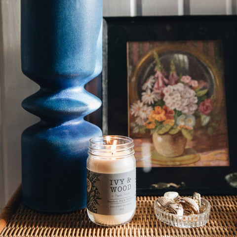 A lit jar candle labeled 'IVY & WOOD' sits on a woven tray beside a large blue vase and a framed floral painting.