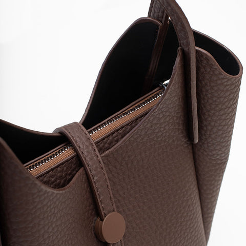 A close-up of a dark brown textured leather tote bag with a top zipper and a matching brown snap button detail, set against a plain white background.