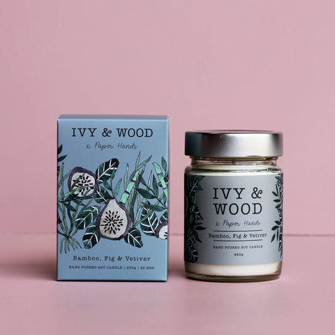 A hand-poured soy candle by Ivy & Wood in the Bamboo, Fig & Vetiver scent, shown next to its light blue illustrated packaging box on a solid pink background.