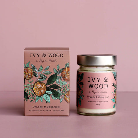 A hand-poured soy candle by Ivy & Wood in the Orange & Ciderleaf scent, shown in its illustrated pink packaging box next to the glass jar candle with a silver lid, set against a solid dusty pink background.