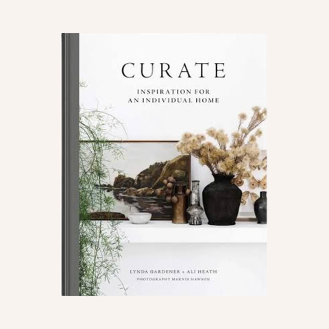 A book titled "CURATE: INSPIRATION FOR AN INDIVIDUAL HOME" shown upright against a light beige background. The cover features a white background with a central styled interior scene, including a landscape painting, dried flowers in a dark vase, and small decorative vessels.