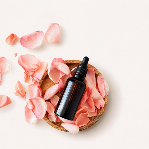 An amber glass dropper bottle with a black cap, styled in a flat-lay arrangement on a light cream background with scattered pink rose petals in a small wooden bowl.