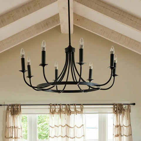 A black multi-light chandelier with candle-style bulbs hanging in a room with a high wood-beamed ceiling and beige curtains.