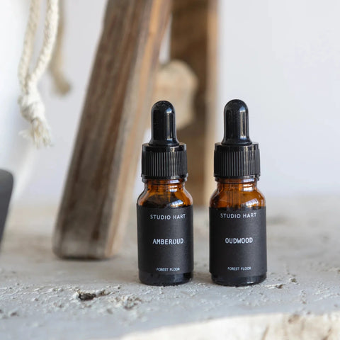 Two amber glass dropper bottles labeled Studio Hart Amberwood and Osduwood, standing on a textured stone surface with a rustic wooden bench in the background.