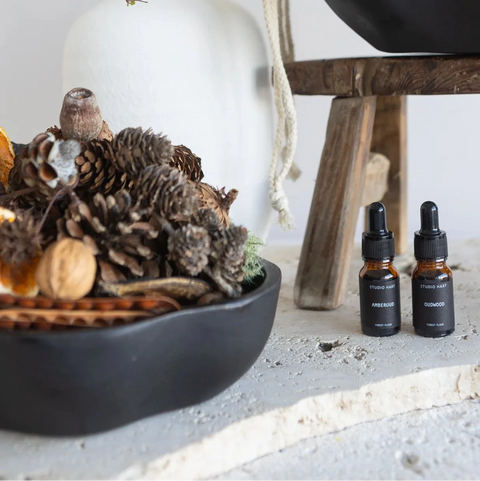 Two small bottles of essential oil on a surface with decorative elements like a bowl of pinecones and branches.