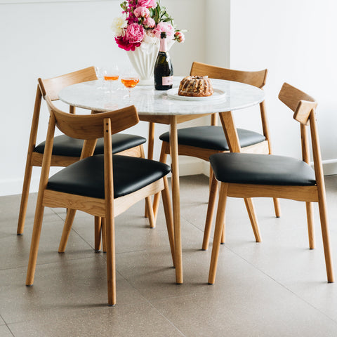 A modern dining set featuring a round white marble-top table and four mid-century style wooden chairs with black upholstered seats, arranged in a bright dining room setting.