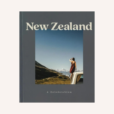 A book cover titled "New Zealand: A Celebration" featuring a dark gray background and a central travel photograph of a person standing on the hood of a vintage car in a scenic New Zealand landscape.