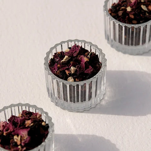 Three small clear ribbed glass cups filled with dried dark purple and brown flower petals, arranged on a white surface with soft shadows.