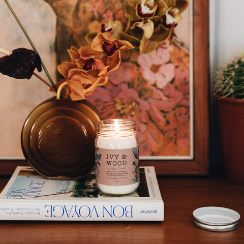 A lit jar candle labeled 'IVY & WOOD' sits on a book on a wooden table, with a vase of flowers and framed wall art in the background. The candle lid rests nearby on the table.