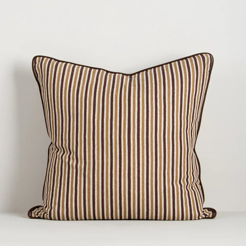 A square throw pillow with vertical stripes in brown, beige, and white, finished with solid brown piped edges, set against a plain light gray background.