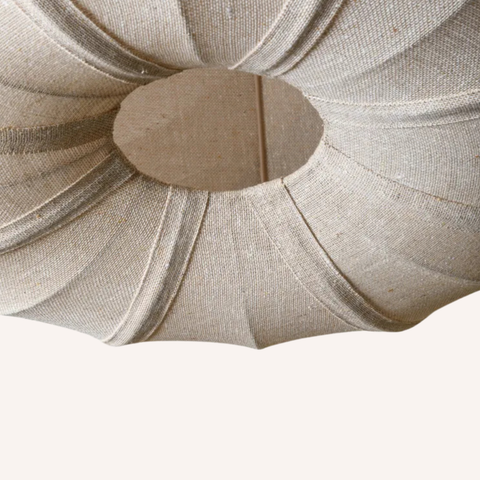 A close-up of a beige woven fabric or jute lampshade with a pleated, flared design and a circular opening at the top, shown against a plain off-white background.