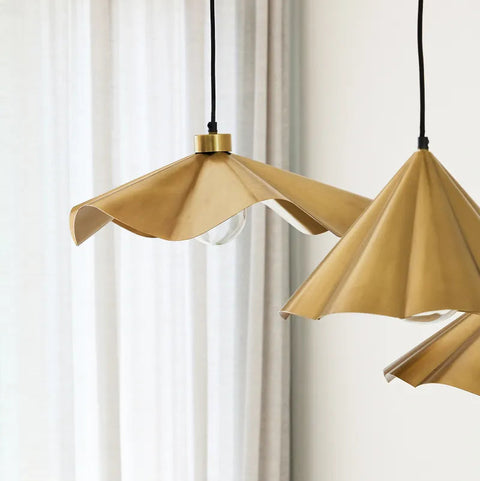 A close-up of two modern pendant lights with pleated, fan-like shades in a warm gold or brass tone, hanging against a soft white curtain and wall.