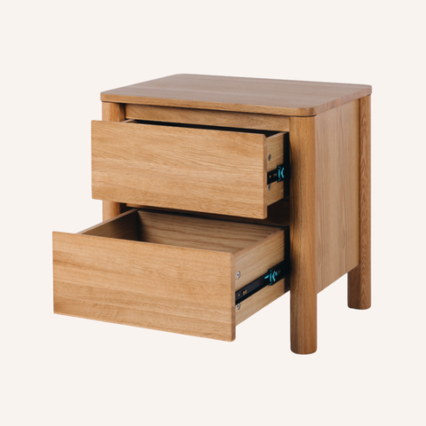 A light oak wood two-drawer nightstand with one drawer pulled open, showing metal drawer hardware, photographed on a solid off-white background.