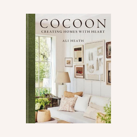 The book cover of 'Cocoon: Creating Homes with Heart' by Ali Heath, featuring a warm, neutral-toned interior photograph of a living room with a sofa, pillows, and framed wall art, set against a light cream background.