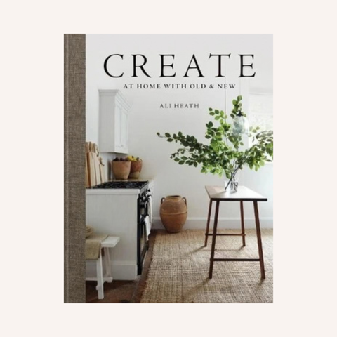The book cover of 'Create At Home With Old & New' by Ali Heath, featuring a warm, rustic kitchen scene with a white stove, a wooden stool holding a vase of greenery, and a woven rug, set against a light beige background.